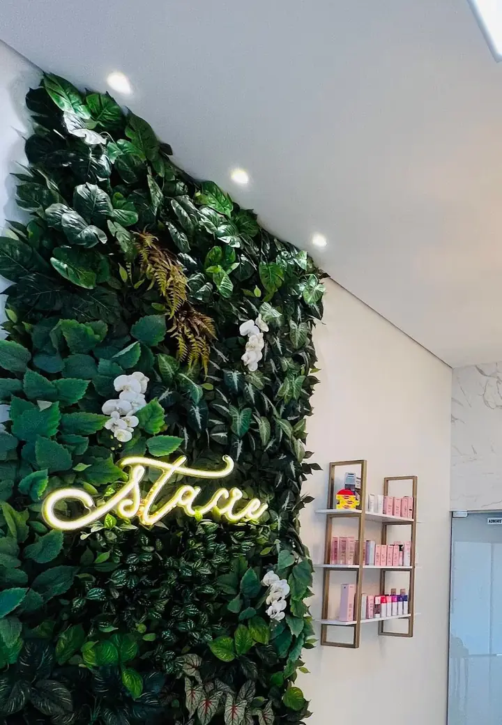 Parede verde com orquídeas, letreiro neon “stacie” e prateleira dourada com produtos